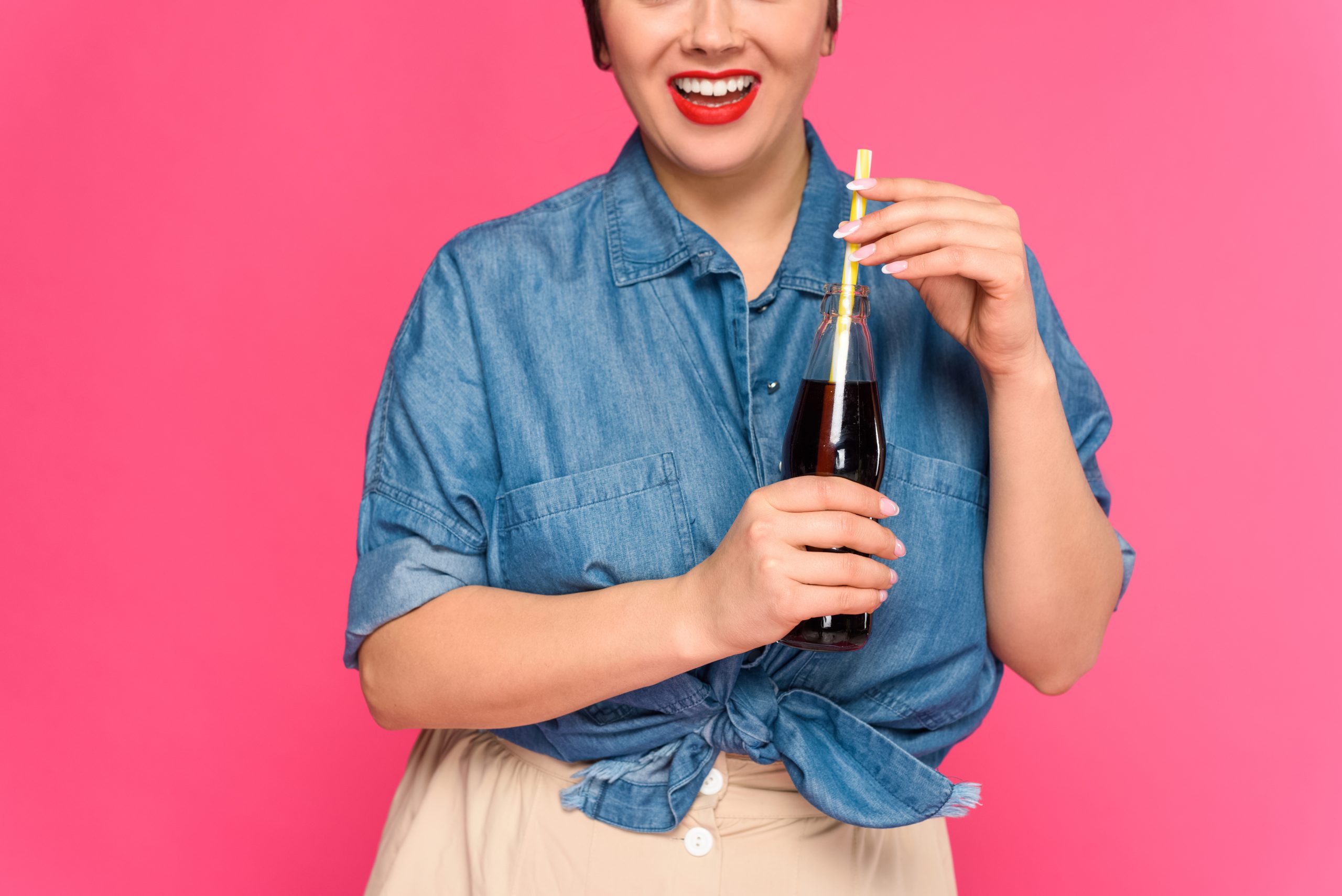 A cropped shot of a smiling woman holding a glass bottle of diet soda with a straw, illustrating the topic of **diet soda on keto**. The vibrant pink background and casual denim outfit emphasize the relaxed and enjoyable aspect of incorporating diet soda into a keto diet.