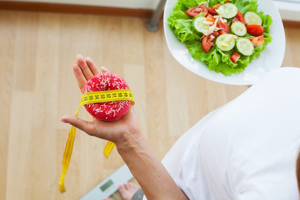 Diet concept, woman measures weight on electronic scales while holding calorie donut.