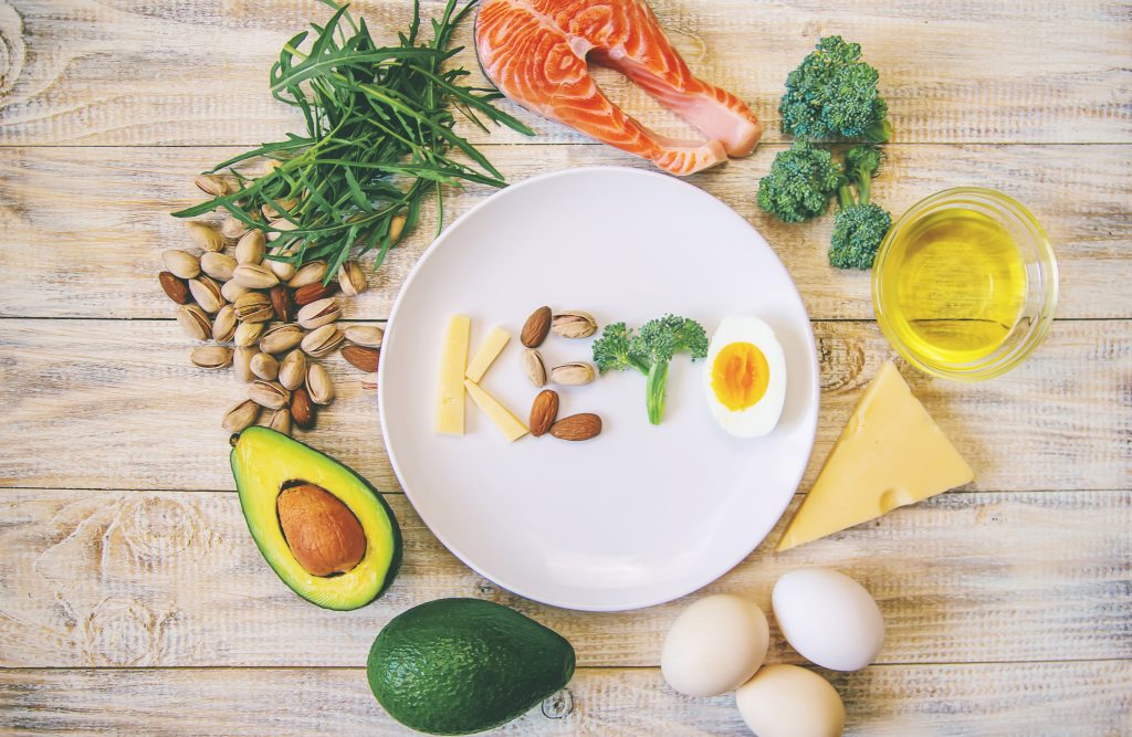 A heart-shaped arrangement of keto-friendly foods including avocado, salmon, broccoli, nuts, cheese, eggs, and olive oil, representing the connection between the keto diet and heart health.