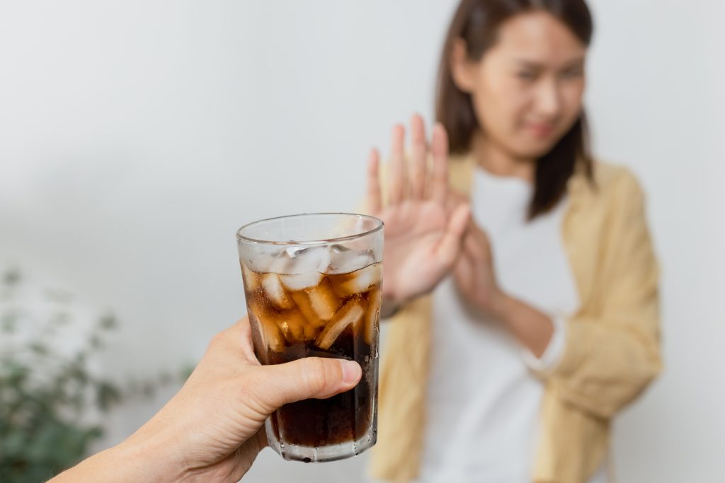 A hand holding a glass of diet soda with ice, while a woman in the background gestures "no" with her hand, illustrating the debate on can you drink diet pepsi on keto. This image captures the concern and considerations regarding the consumption of diet sodas while following a ketogenic diet.