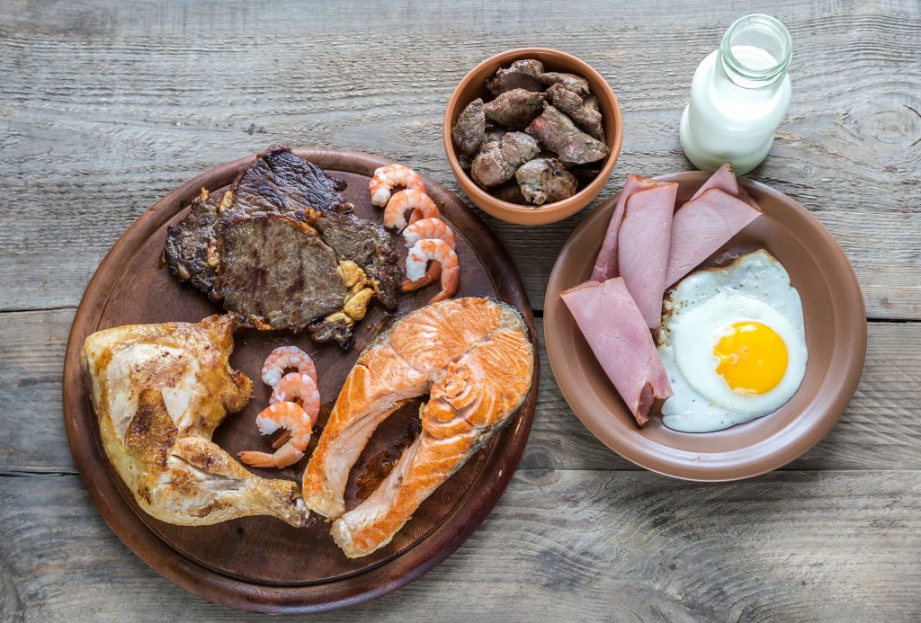 The image showcases a hearty spread of various high-protein foods, ideal for a discussion on the keto diet vs carnivore diet. It features a rich array of animal proteins including grilled steak, roasted chicken leg, a salmon fillet, and shrimp, complemented by sliced ham and a sunny side up egg. Additionally, there is a bowl of cooked liver pieces and a bottle of milk, providing a full spectrum of carnivore and ketogenic diet-friendly options. This selection highlights the focus on meat and high-protein foods typical of both diets, emphasizing their roles in such nutritional plans.