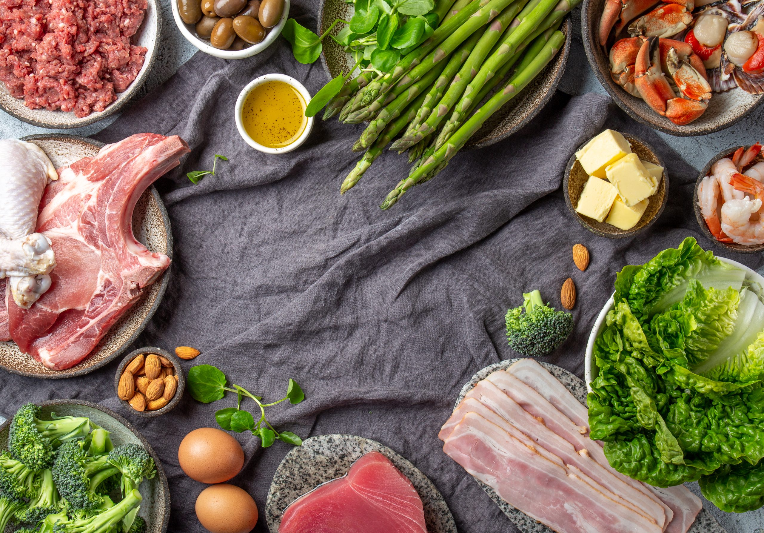 The image presents a variety of foods representing both plant-based and animal-based nutrients, ideal for a comparison of different diet types such as the carnivore diet vs keto diet. Visible in the arrangement are meats like beef, chicken, and pork, seafood such as shrimp and crab, alongside dairy products like butter. Complementing these are plant-based items including asparagus, almonds, broccoli, and leafy greens, providing a rich contrast of dietary options. This setup effectively illustrates the diverse sources of protein, carbohydrates, and fats available in different diets.