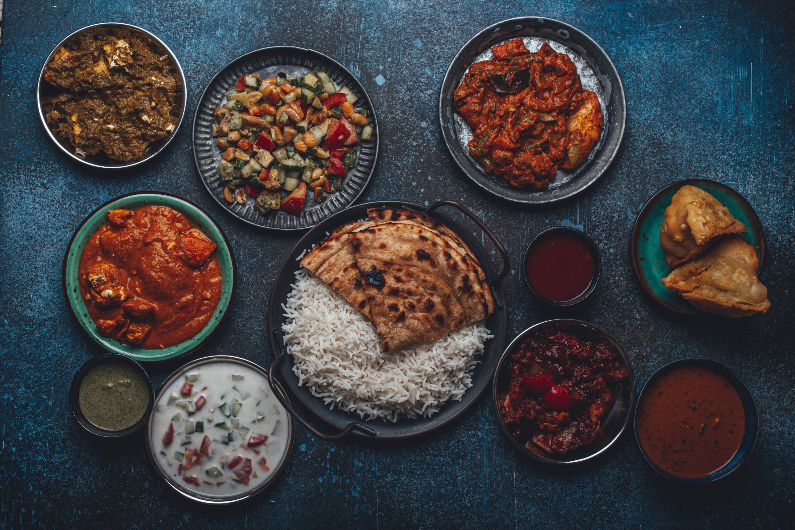 Assorted Indian ethnic food buffet on rustic concrete table from above: curry, fried samosa, rice biryani, dal, paneer, chapatti, naan, chicken tikka masala, traditional dishes of India for dinner. Ayurvedic Detox for Weight Loss concept