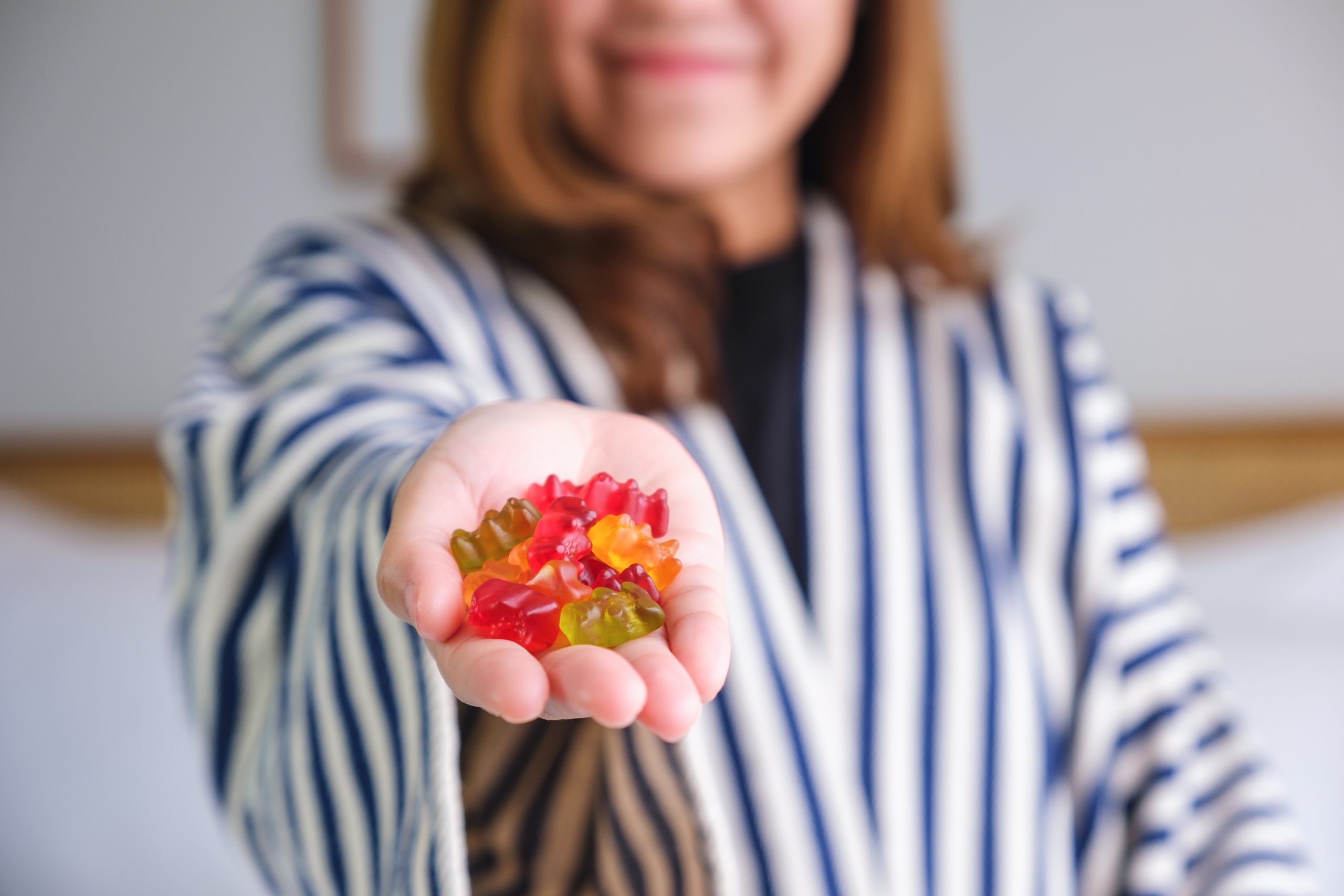 A young woman in a striped blazer smiling and holding a handful of colorful keto diet gummies directly towards the camera, suggesting a health-conscious alternative to traditional sweets.