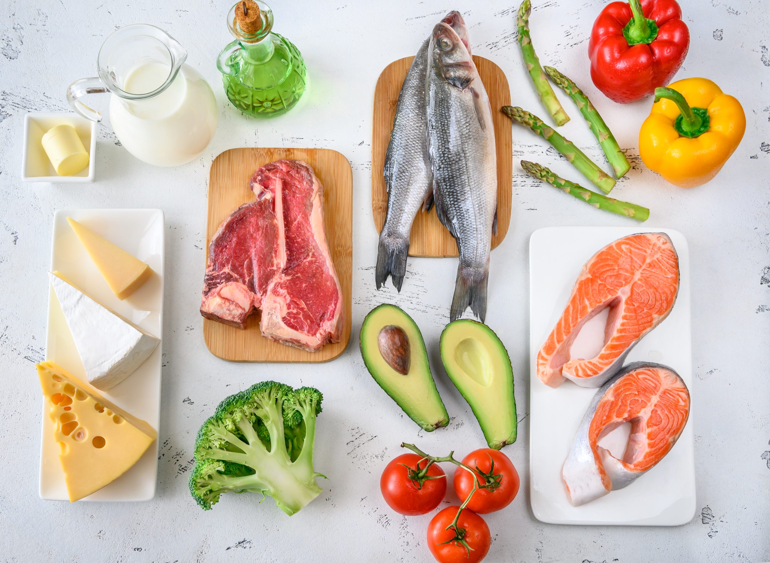 Image of various foods suitable for the Modified Atkins Diet, including a selection of healthy fats, proteins, and low-carb vegetables. Featured items are raw steaks, whole fish, salmon fillets, avocado, broccoli, tomatoes, bell peppers, asparagus, assorted cheeses, a jug of milk, and bottles of oil.