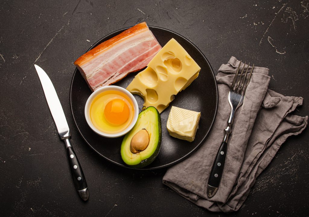 Keto diet foods on a black plate, including bacon, cheese, a raw egg, avocado, and butter, representing what is dirty keto diet.