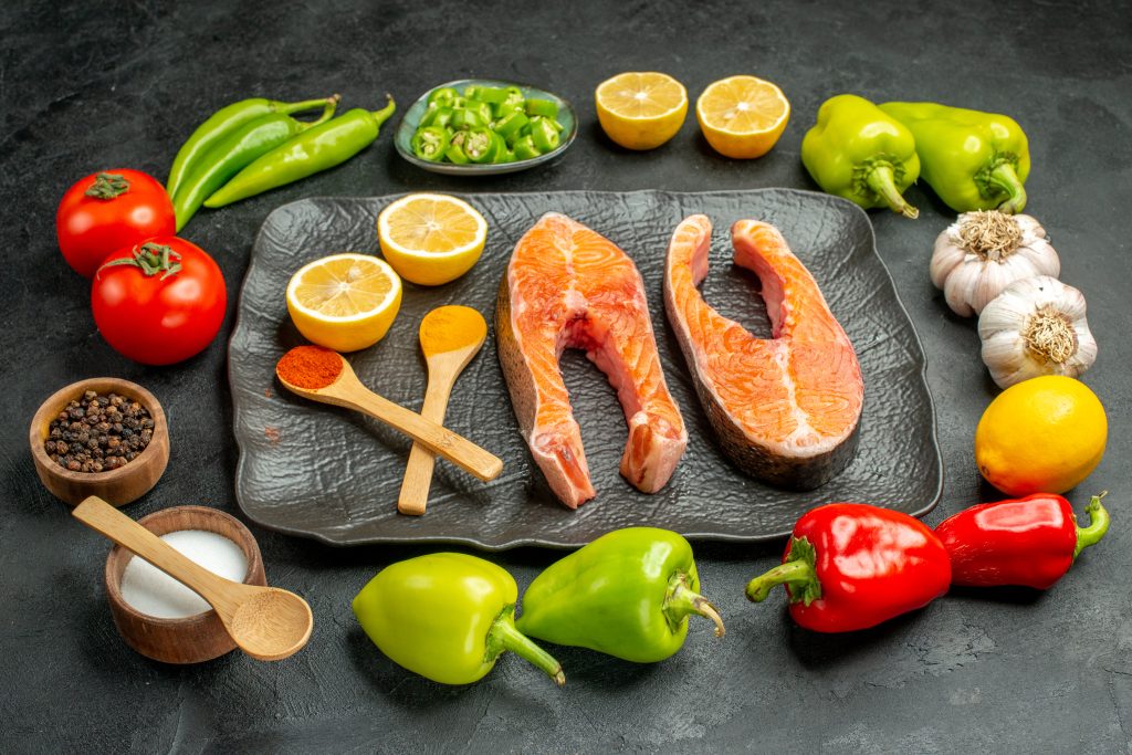 The image depicts a variety of fresh ingredients suitable for both the Atkins Diet and Keto Diet. Included are salmon steaks, bell peppers, tomatoes, lemons, garlic, and various spices, showcasing foods that align with low-carb, high-protein dietary plans, highlighting the atkins vs keto diet differences.