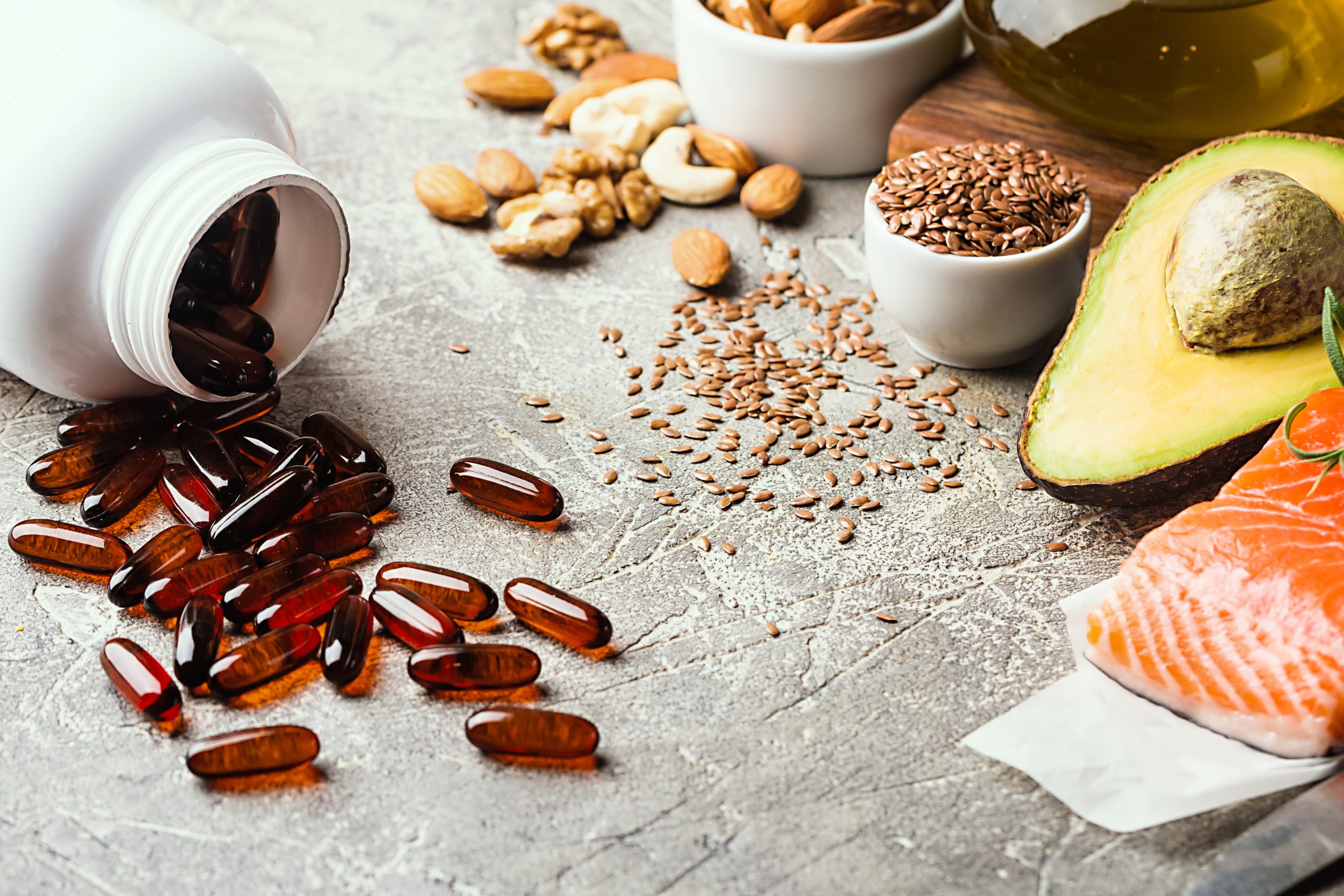 A variety of healthy fats and keto diet supplements displayed on a textured surface, featuring a spilled bottle of dark red keto diet pills, alongside almonds, walnuts, flaxseeds in white bowls, avocado, salmon, and a bottle of olive oil, illustrating components of a nutritious ketogenic diet.