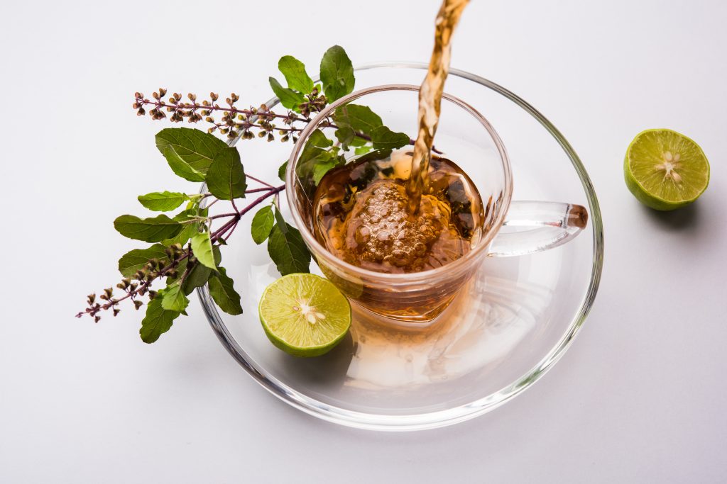 a glass cup of herbal tea being poured, garnished with fresh basil leaves and accompanied by halved lime, displayed on a clear saucer.