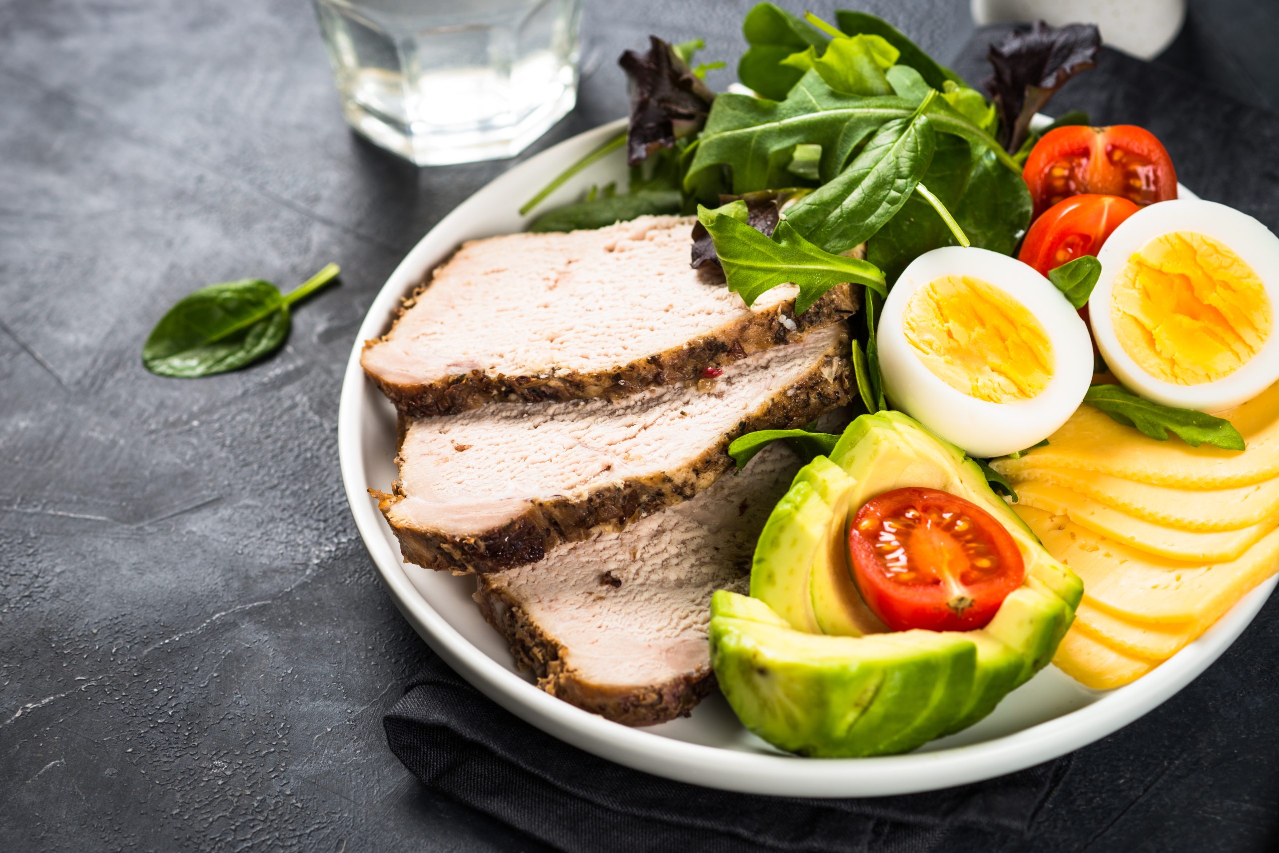 Healthy meal for South Beach Diet vs Keto featuring sliced turkey, boiled eggs, avocado, cheese, and leafy greens on a white plate against a dark background.