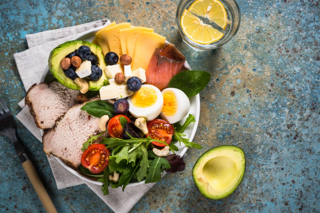 A nutritious low-carb meal featuring avocado, boiled eggs, cheese, nuts, blueberries, cherry tomatoes, leafy greens, and sliced meat, accompanied by a glass of lemon water, perfect for an Atkins diet plan menu.