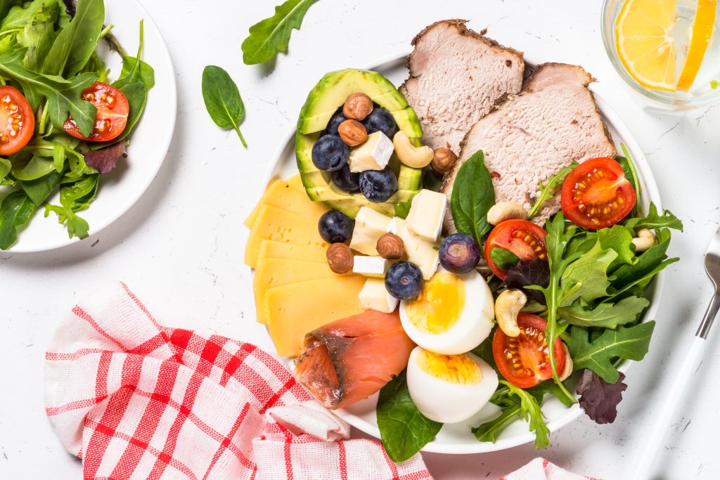 Healthy meal for South Beach Diet Phase 2 featuring sliced turkey, avocado, cheese, nuts, boiled eggs, leafy greens, and tomatoes arranged on a white plate with a red-checkered napkin.