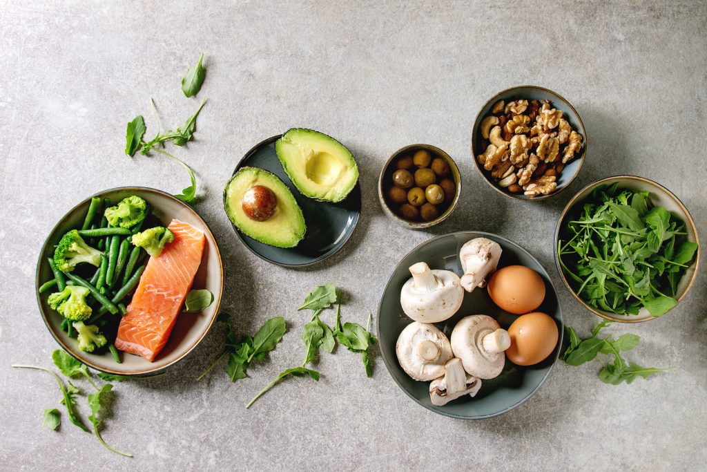 Ingredients for South Beach Diet Phase 1 meal, including salmon, avocado, broccoli, green beans, nuts, olives, eggs, mushrooms, and fresh greens, arranged on a gray background.