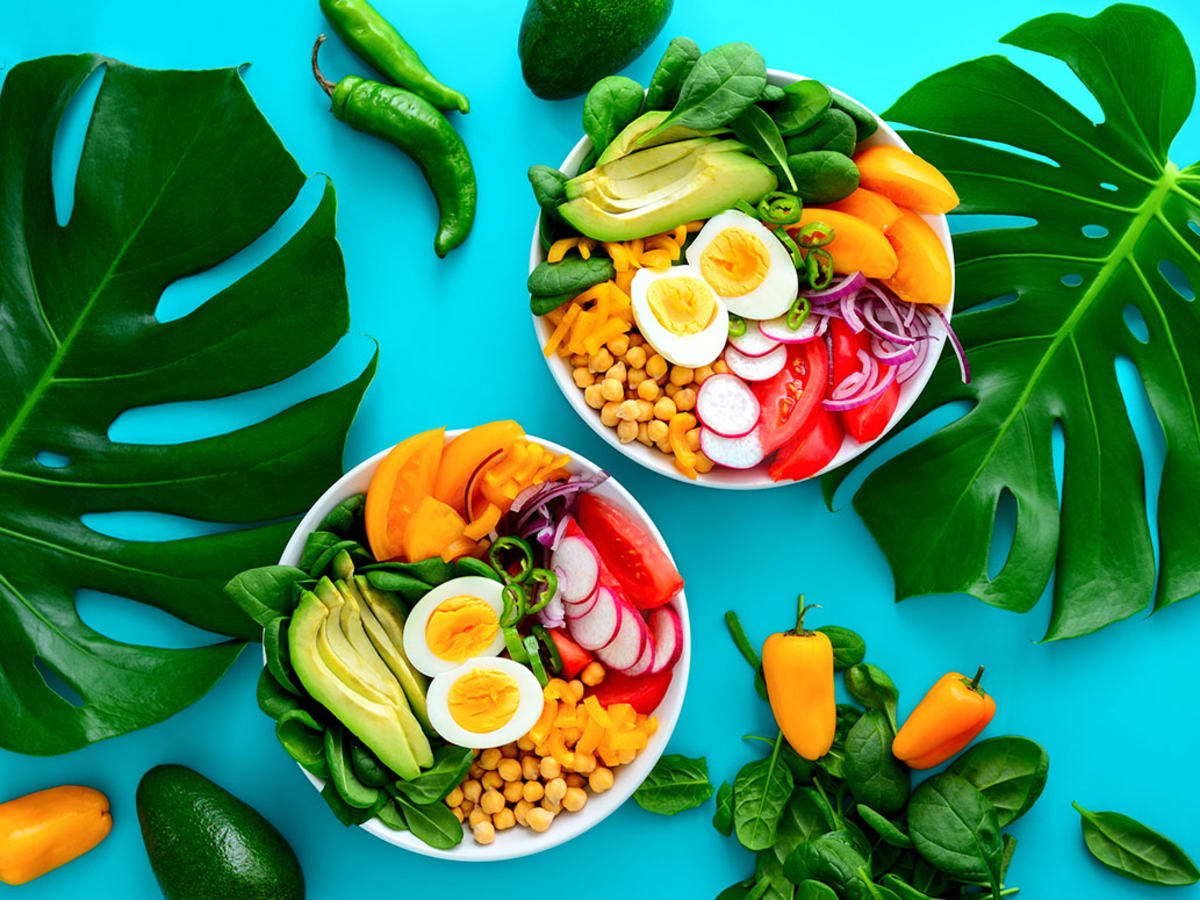 Colorful South Beach Diet Phase 1 meal featuring fresh vegetables, boiled eggs, avocados, and leafy greens arranged in bowls on a vibrant blue background with tropical leaves.