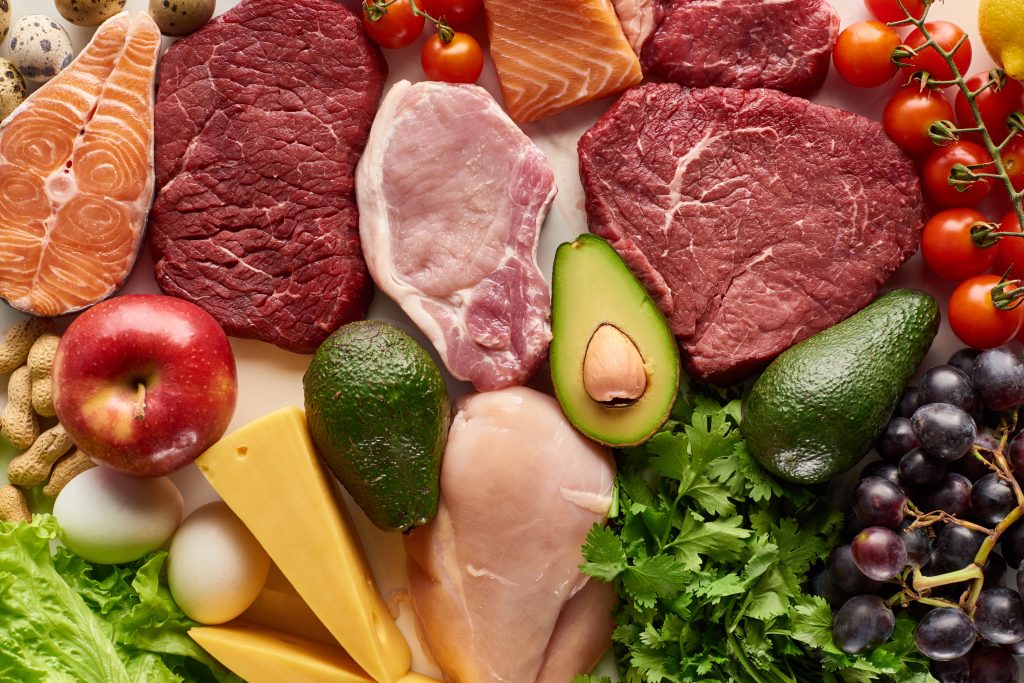 Top view of assorted raw meat, poultry, fish, eggs, vegetables, fruits, nuts, greenery and cheese