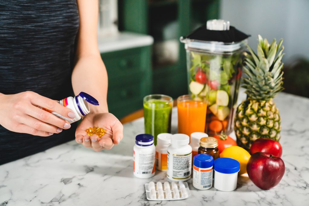 An array of dietary supplements and healthy foods on a kitchen counter, highlighting a woman choosing omega-3 capsules, potentially akin to keto diet pills, surrounded by fresh fruits like pineapple and apples, and green vegetable juices, illustrating a health-conscious lifestyle.