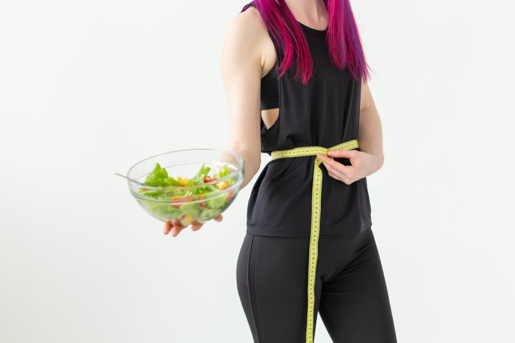 Young unidentified woman athlete with purple hair measures the waist of a measuring tape and holds a vegetable salad. Concept of proper nutrition and good shape